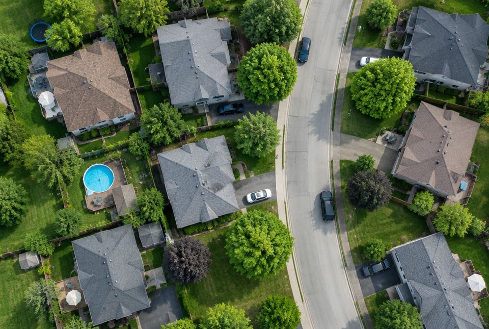 A view from high above a neighbourhood showing house roofs, trees, a curving street, cars and a backyard pool.