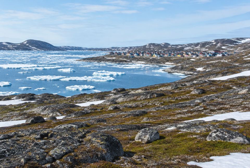 A small Arctic town of colourful houses beside a bay with floating ice, seen across rocky land with patches of snow.