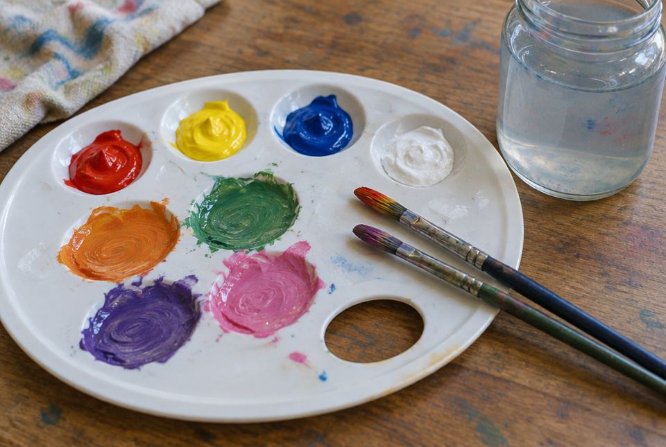 A paint palette with red, yellow, blue and white paint, and mixed orange, green, violet and pink paint, with two brushes and a jar of water.