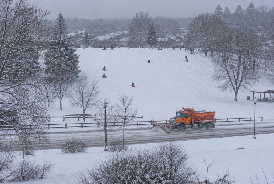A snowplow clearing a road while people go sledding on a snowy hill in the background.