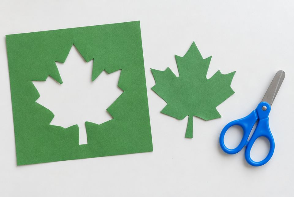 A green paper square with a maple-leaf-shaped hole beside the green paper leaf that was cut out of it, and a pair of scissors.