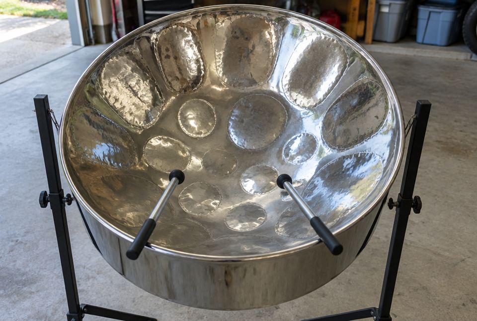A shiny steelpan drum on a stand with two sticks, in a garage.