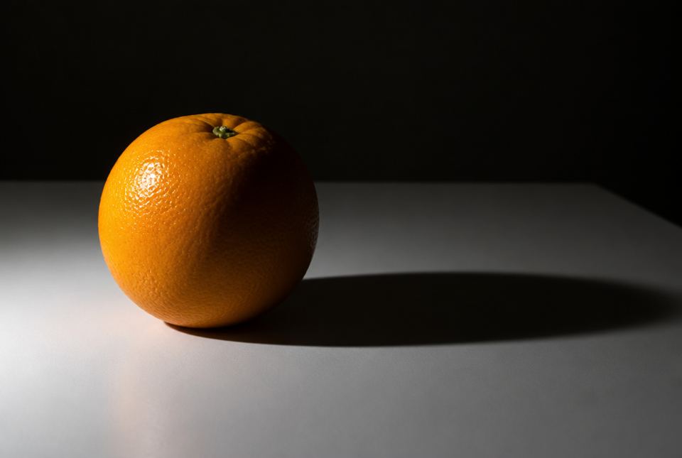 An orange on a white table, lit from the left, with its right side in shadow and a long dark shadow stretching to the right.