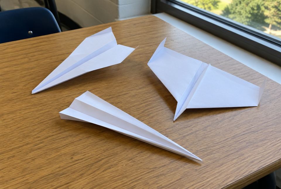 Three white paper airplanes with different wing shapes resting on a classroom table by a window.