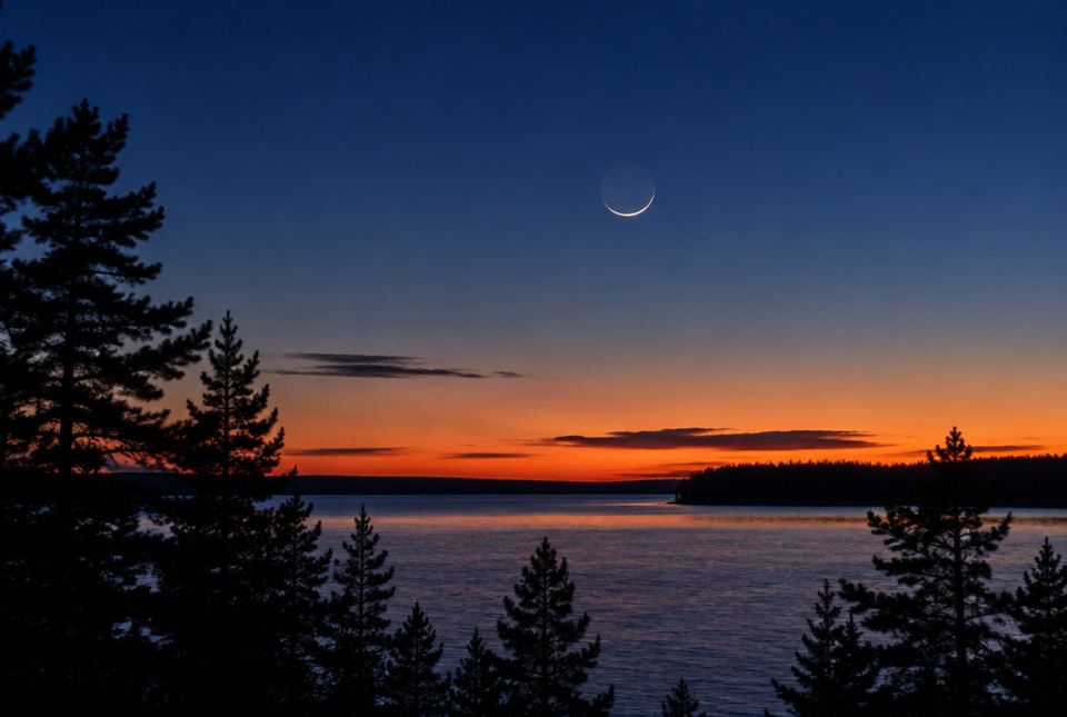 A thin crescent Moon in the twilight sky above a lake and pine trees.
