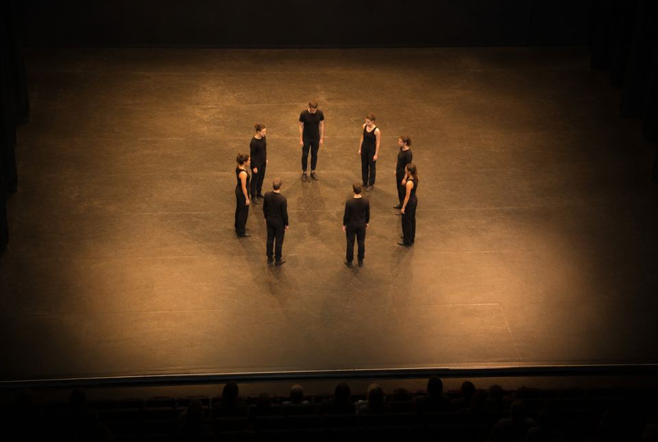 A group of dancers in black standing in a circle on a stage, seen from above.
