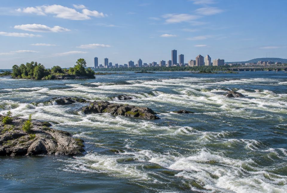 White-water rapids flowing over rocks in a wide river, with a city skyline in the distance.