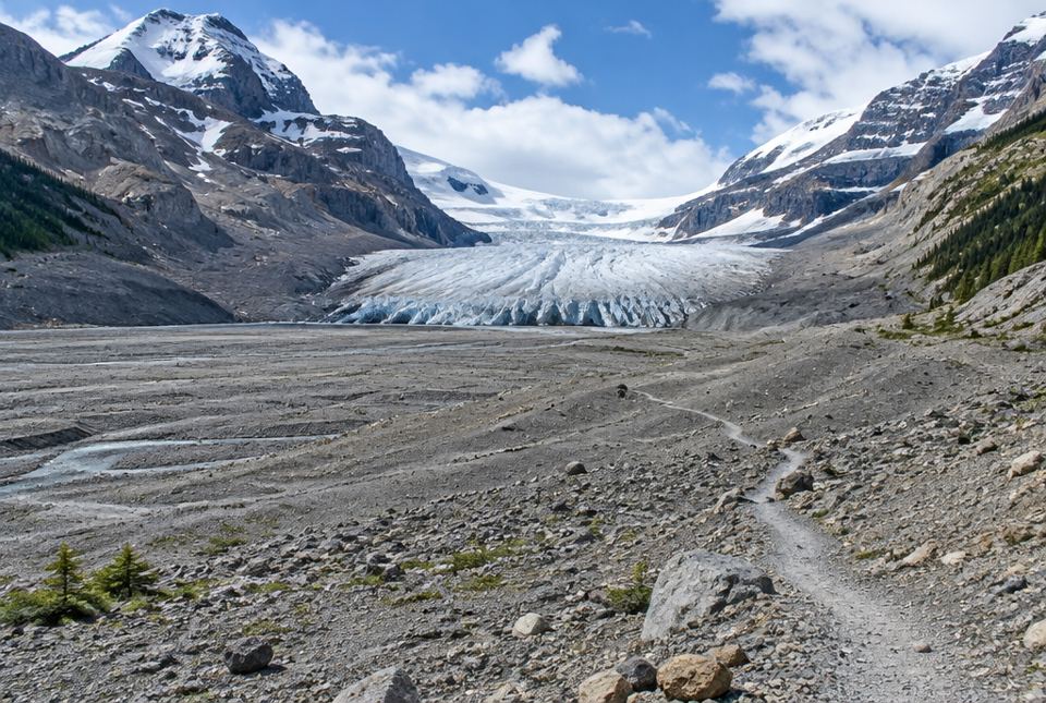 A glacier's icy edge fills a mountain valley, with a long stretch of bare grey rock and a trail in front of it.