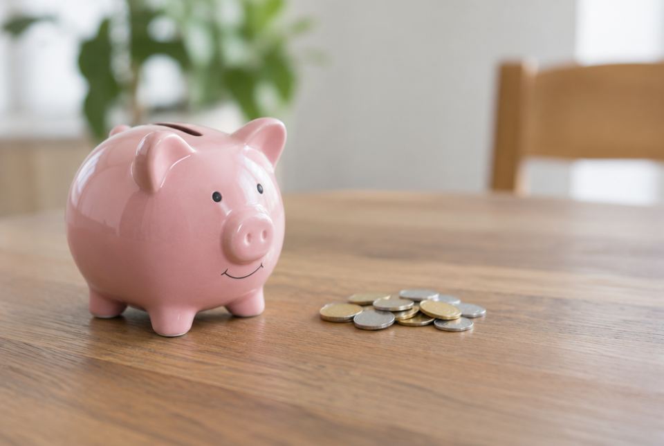 A pink piggy bank sits on a wooden table next to a small pile of silver and gold coloured coins.