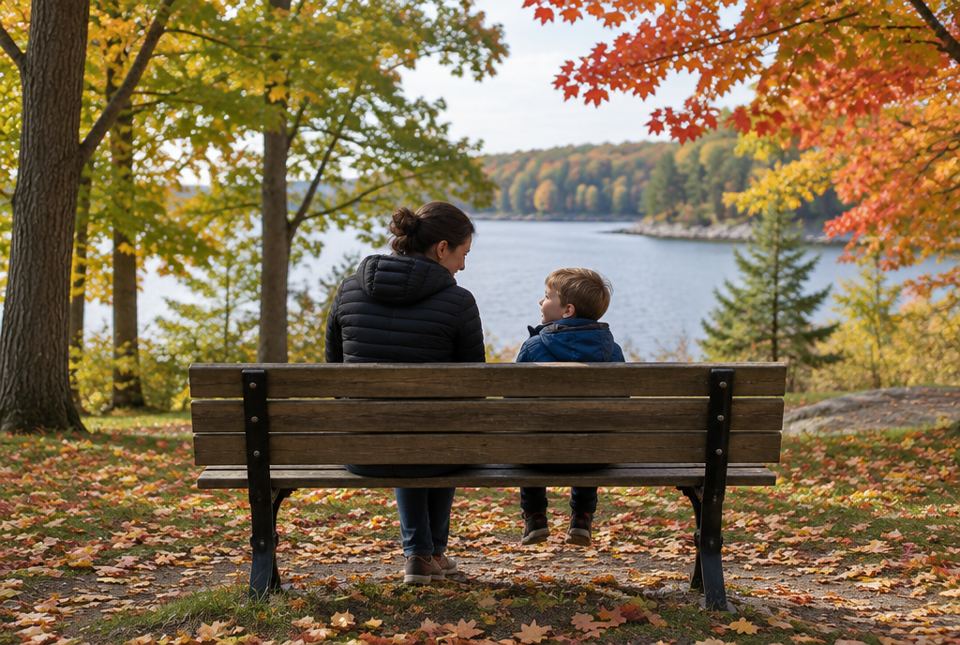 A grown-up and a child sit on a park bench under autumn maple trees, turned toward each other as they talk.
